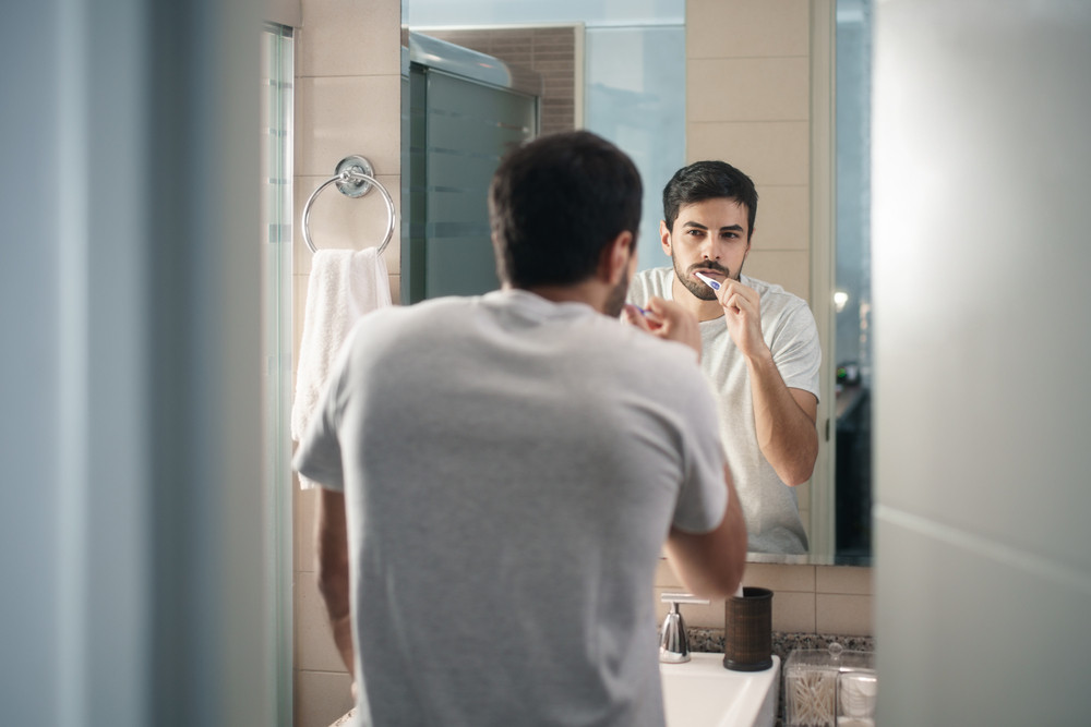 Hispanic person with beard grooming in bathroom at home for morning routine and body care. White metrosexual man brushing teeth in front of mirror